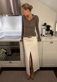 Καφέ μακρυμάνικο πόλο πουκάμισο, λευκή μέγιστη φούστα με μπροστινή σχισμή και ακατέργαστο τελείωμα, ταμπά μπότες με μυτερή μύτη μέχρι τον αστράγαλο, σύγχρονη κουζίνα στο φόντο.