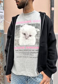Homme portant des écouteurs, un sweat à capuche noir et un T-shirt gris montrant une affiche d'un chaton blanc disparu avec une récompense de 1000 $.