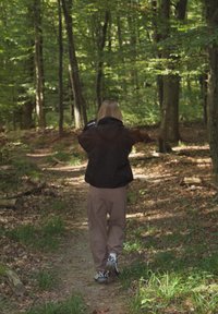 Cilvēks staigā pa meža taku, ģērbies melnā jakā un gaiši brūnās biksēs, ar sporta apaviem. Fona attēlo zaļus kokus un lapotni.