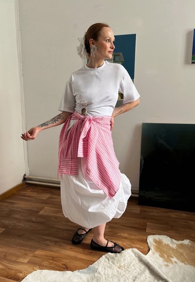 Camiseta blanca con detalle de nudos, superpuesta con un envoltorio a rayas rosa y blanca atado a la cintura, combinada con una falda larga blanca y zapatos planos negros.
