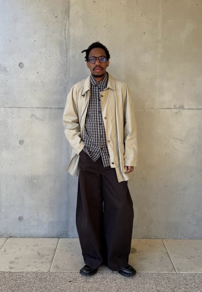 Hombre con gafas que lleva un abrigo beige, camisa a cuadros, pantalones negros anchos y zapatos negros, de pie junto a una pared de hormigón.