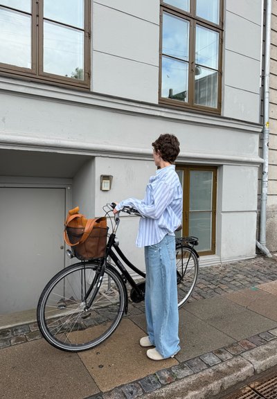 Persona con una camiseta de rayas y vaqueros está de pie en una acera sosteniendo una bicicleta negra con una bolsa marrón en la cesta delantera cerca de un edificio.