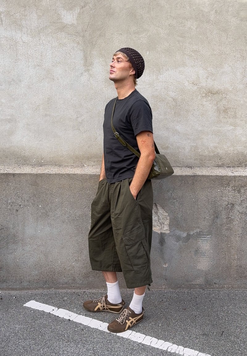 Man som bär en svart t-shirt, gröna vida byxor, bruna sneakers och en svart stickad mössa, står med händerna i fickorna mot en grå vägg.