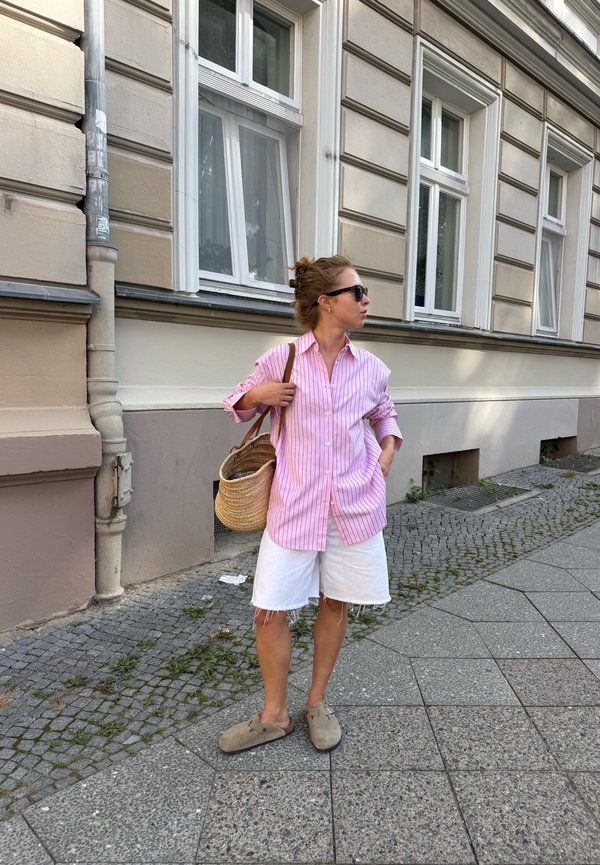 Femme portant des lunettes de soleil, une chemise rose à rayures, un short blanc longueur genou et des chaussures beiges, debout sur un trottoir près d’un bâtiment beige, tenant un sac en paille.