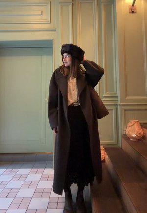 Mujer con abrigo largo oscuro, sombrero de piel negro, suéter estampado y falda negra, de pie en el interior cerca de escalones con linternas encendidas.