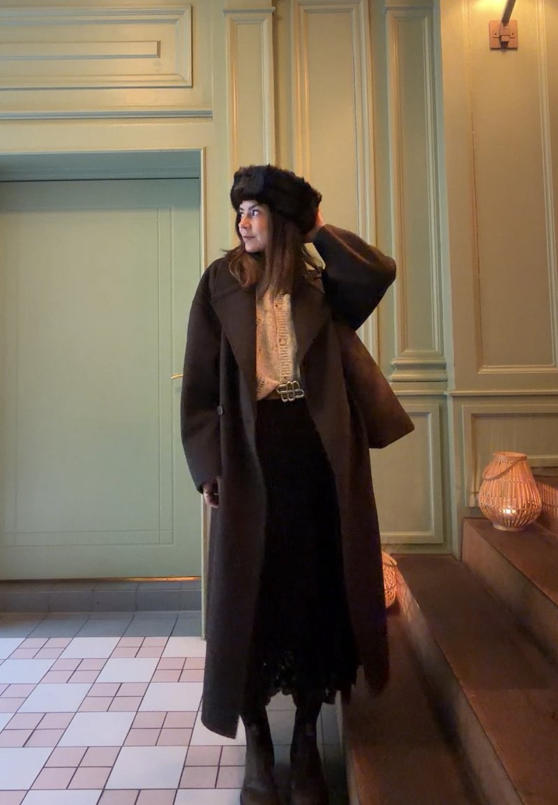 Donna con cappotto lungo scuro, cappello di pelliccia nero, maglione a motivi e gonna nera, in piedi all'interno vicino a scalini con lanterne accese.