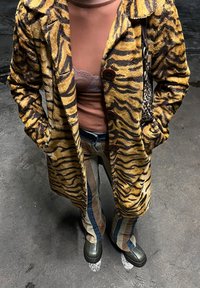 Άτομο που φοράει παλτό με κίτρινες και μαύρες ρίγες τίγρης, καφέ τοπ με δαντελένια λεπτομέρεια, παντελόνι πατς, πράσινα παπούτσ ια, στέκεται σε άσφαλτο.