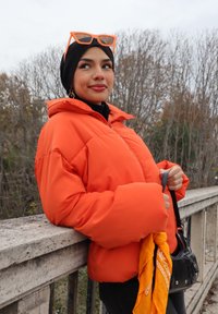 Veste orange gonflée avec une capuche, associée à un hijab noir et des lunettes de soleil orange. Tenant un sac noir et un bandana jaune.