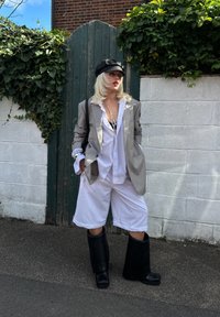 Šedý oversized blejzr, bílá košile na knoflíky, bílé šortky a černé vysoké boty nad kolena; doplněno černou kulichovou čepicí.