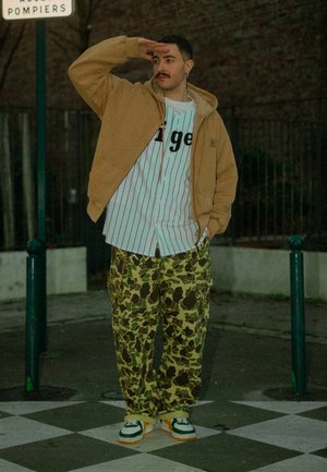 Joven de pie sobre un pavimento a cuadros, vestido con pantalones de camuflaje, camisa a rayas, chaqueta marrón clara y zapatillas deportivas, cubriéndose los ojos con una mano.