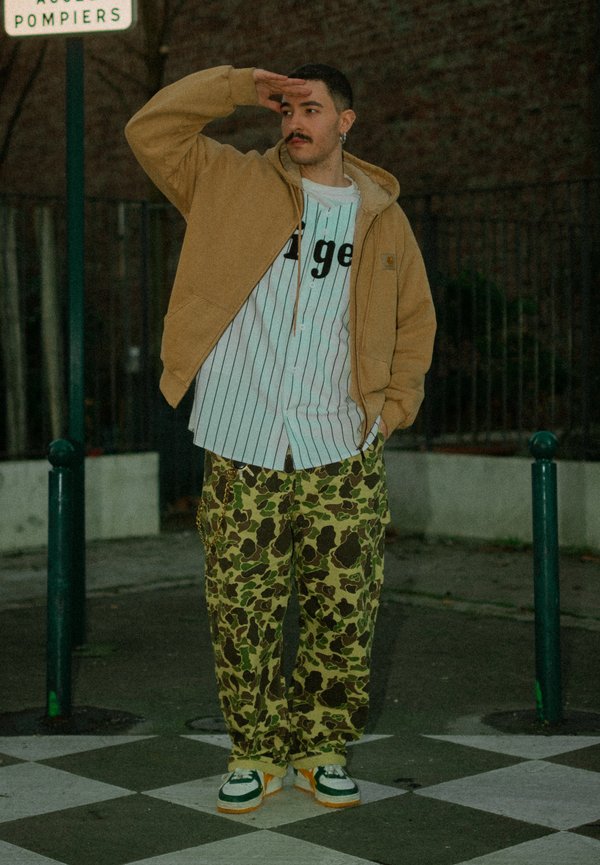 Ung mand står på ternet fortov iført camouflagebukser, stribet skjorte, beige jakke og sneakers, skygger øjnene med én hånd.