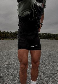 Czarne sportowe spodenki Nike z szarymi akcentami, dopasowanym krojem i białym logo. Osoba ma na sobie czarną koszulkę i zielony plecak.