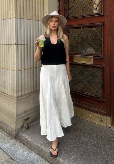 Top sin mangas negro, falda blanca fluida, sombrero de ala ancha color tostado, sosteniendo una bebida verde, de pie cerca de una puerta de madera con detalles decorativos.