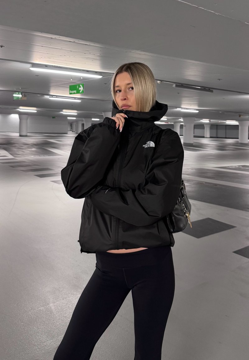 Woman with blonde hair wearing a black North Face jacket and black leggings, standing in a mostly empty, well-lit parking garage.
