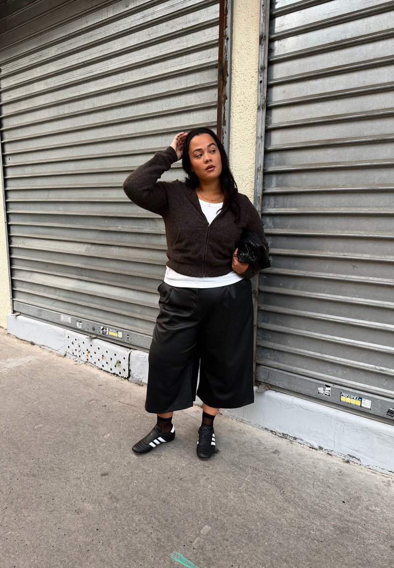 Svart kort genser, hvit skjorte, svarte vide bukser i kunstskinn og svarte sportssko med hvite striper. Står mot en metallrulleskodde.