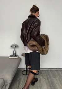 Veste en cuir marron avec une coupe oversize, portée sur une chemise blanche à col. Tient un châle en fourrure beige, un short noir et des collants transparents avec des bandes aux chevilles.