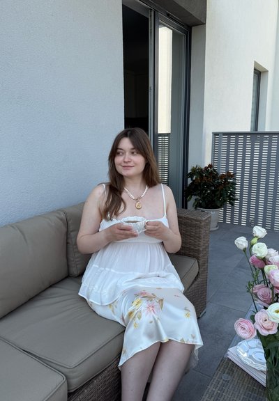 Vestido blanco floral con un diseño fluido, sentada en un sofá beige. Sosteniendo una taza de té blanca, rodeada de un ramo de rosas rosadas y blancas.