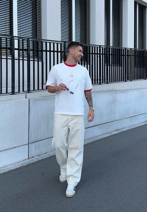 Camiseta blanca con cuello rojo, pantalones cremosos informales y zapatillas blancas. Sosteniendo unas gafas de sol, de pie sobre una superficie pavimentada cerca de un edificio.