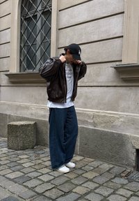Veste bomber en cuir marron portée sur un t-shirt blanc avec texte, associée à un pantalon large en denim bleu et des baskets blanches, debout sur une rue pavée.