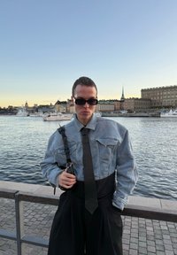 Džinsinis švarkas su sutrumpintu dizainu, juoda kaklaraištis ir platūs juodi kelnių; su akinių nuo saulės derinys; vandens prieigos fono su pastatais.