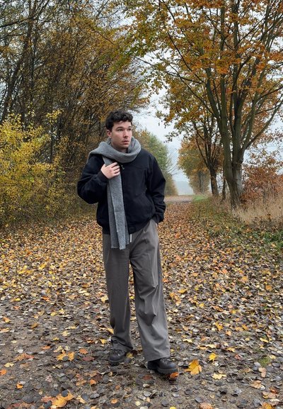 Cazadora bomber negra, bufanda gris, pantalones beige de pierna ancha y zapatos oscuros. La escena muestra un camino bordeado de follaje otoñal y árboles.
