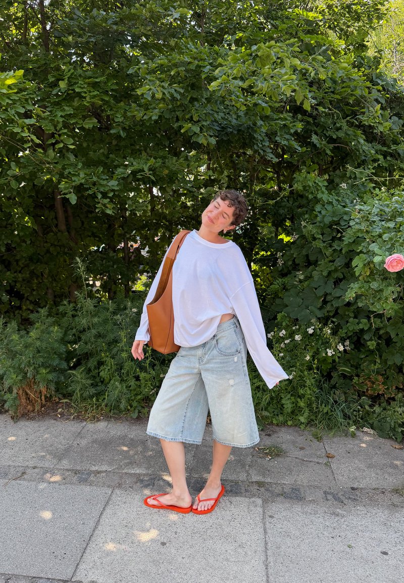 Personne debout sur un trottoir devant des buissons verts, portant une chemise blanche à manches longues, un short en denim, des tongs rouges et portant un sac marron.