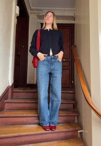 Γυναίκα με μαύρη ζακέτα, μπλε τζιν και κόκκινα παπούτσια στέκεται σε ξύλινη σκάλα εσωτερικού χώρου με τα χέρια στις τσέπες και κόκκινη τσάντα στον ώμο.