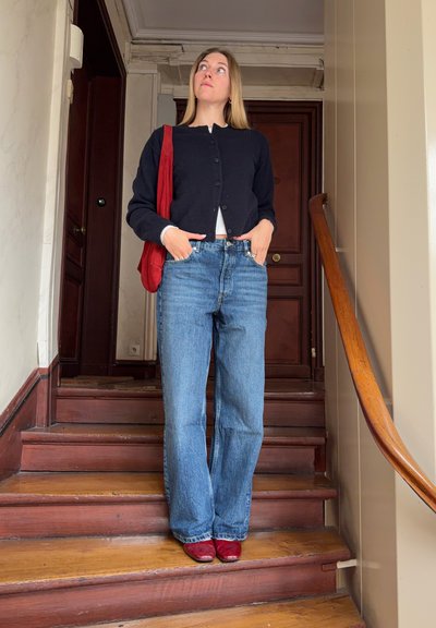 Mujer con cárdigan negro, vaqueros azules y zapatos rojos, de pie en unas escaleras de madera dentro de una casa, con las manos en los bolsillos y un bolso rojo al hombro.