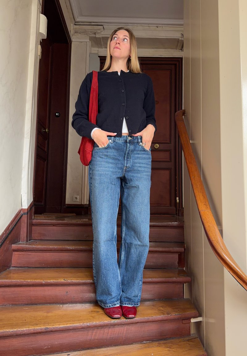 Vrouw die een zwart vest draagt, blauwe jeans en rode schoenen, staande op houten trappen binnen met handen in de zakken en een rode tas over de schouder.
