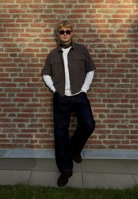 Kurzärmliges braunes Hemd mit Knöpfen, darunter ein langärmliges weißes Hemd, blaue Jeans, braune Wildlederschuhe, Sonnenbrille, an einer Backsteinmauer stehend.