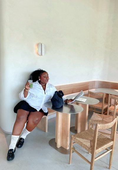 Una persona sentada sostiene una bebida verde, vistiendo una camisa blanca de botones y una falda negra. A su alrededor: mesas de metal, sillas de madera y paredes en tonos neutros.