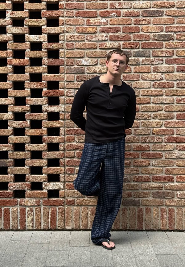 Homme avec des lunettes portant une chemise noire à manches longues, un pantalon à carreaux et des tongs, appuyé contre un mur en briques avec une jambe levée.