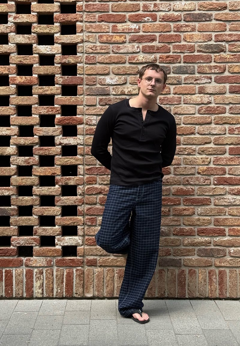 Homme avec des lunettes portant une chemise noire à manches longues, un pantalon à carreaux et des tongs, appuyé contre un mur en briques avec une jambe levée.
