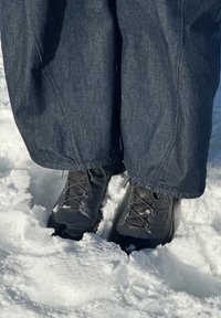 Personne portant un pantalon foncé et des bottes d'hiver noires, debout dans la neige avec de la neige collée aux bottes et aux ourlets du pantalon.