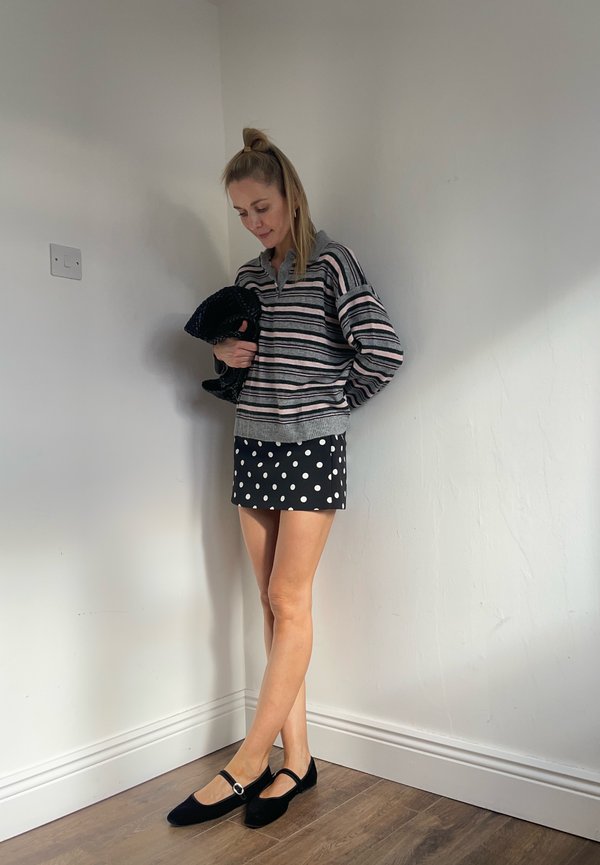 Woman standing indoors against a plain wall wearing a striped sweater, polka dot skirt, black Mary Jane shoes, and holding a textured black clutch.