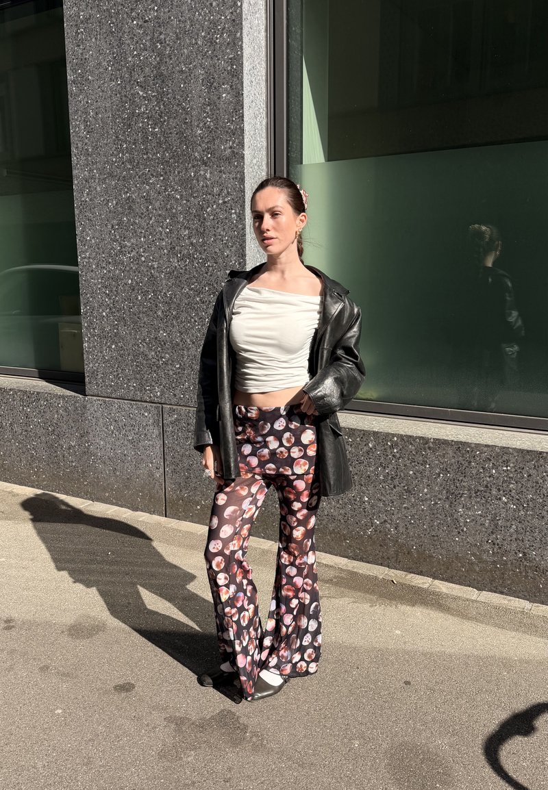 Femme debout à l'extérieur à côté d'un mur sombre tacheté portant un haut blanc froncé, une veste en cuir noire et un pantalon évasé avec un motif circulaire coloré.