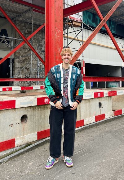 Chaqueta bomber acolchada con detalles en verde y naranja, camiseta gráfica, pantalones negros holgados y zapatillas multicolor, de pie frente a un fondo de construcción.