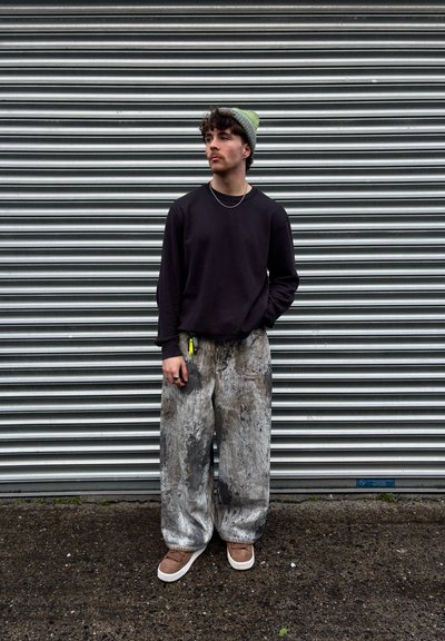 Modelo que lleva una camisa de manga larga negra texturizada y unos pantalones gris grandes con un patrón, complementados con zapatos marrones y un gorro verde.