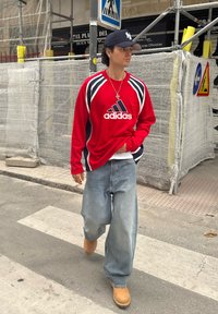 Mladý muž v červenej mikine Adidas, modrej šiltovke, voľných džínsach a béžových topánkach kráča po mestskej ulici pri stavebných zábranách.