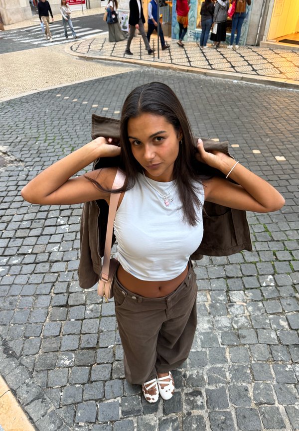 Mujer joven en calle adoquinada ajustándose la chaqueta sobre un crop top blanco, lleva pantalones marrones y zapatos con estampado de vaca, con peatones al fondo.