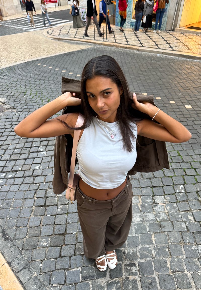 Jeune femme dans une rue pavée ajustant sa veste sur un crop top blanc, portant un pantalon marron et des chaussures à imprimé vache, avec des piétons en arrière-plan.