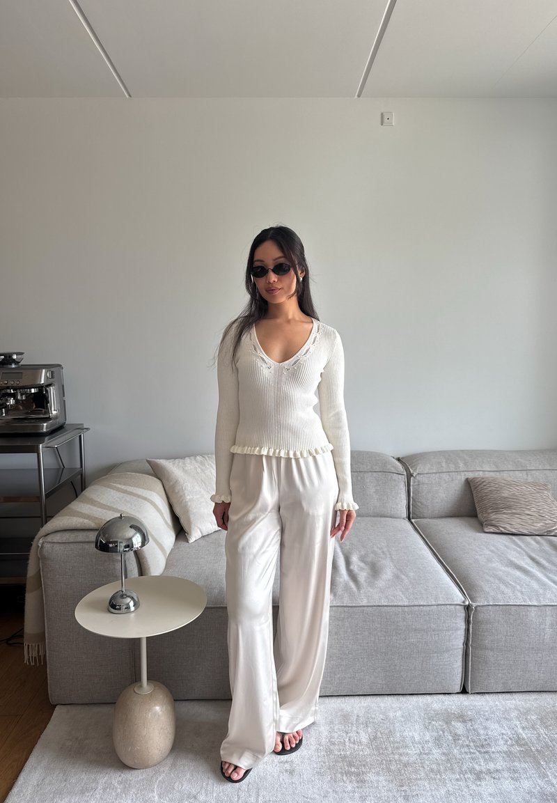 Femme aux longs cheveux foncés et lunettes de soleil, debout à l'intérieur près d'un canapé gris, portant un pull crème et un pantalon en soie, à côté d'une table d'appoint ronde.