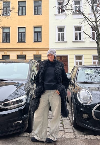 Abrigo negro de imitación piel con mangas texturizadas, suéter de cuello alto negro, pantalones amplios gris claro y gorro gris con diseño. Dos coches aparcados en el fondo.