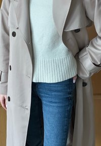 Bézs színű trench coat egy világosszürke, bordás szegélyű pulóver fölött, kék farmernadrággal párosítva. A kabáton nagy gombok találhatók, és laza szabású.