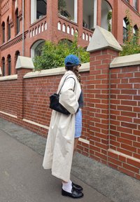 Beige lang frakk, mørk genser, lys blå stripete shorts, svart håndveske, og loafers med hvite sokker. Bakgrunnen har murvegg og grønnskjær.