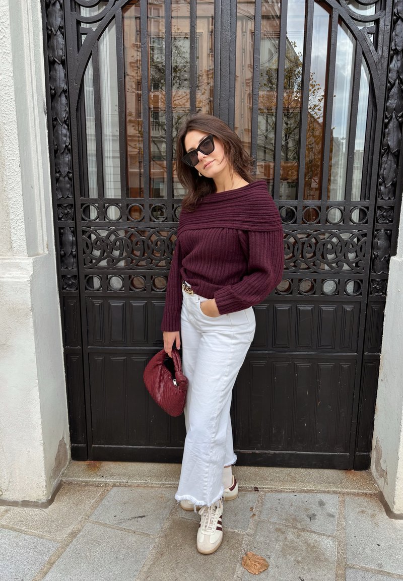 Bordeauxroter, gerippter Pullover, weiße Schlagjeans mit ausgefranstem Saum, cremefarbene Sneakers mit roten Streifen und eine burgunderrote, gesteppte Handtasche. Schwarzer Eisentor-Hintergrund.