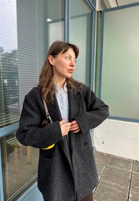 Μαύρο παλτό σε σχέδιο καρέ με φαρδιά γραμμή, υφασμάτινη υφή, με κοψίματα στις λαιμόκοψεις και δύο μπροστινές τσέπες. Συνδυασμένο με πουκάμισο με ρίγες σε ανοιχτό μπλε χρώμα.