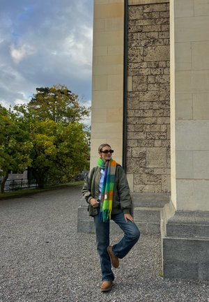 Chaqueta bomber verde, camisa gris, jeans azules y botas marrones. Bufanda multicolor con rayas en verde, azul, amarillo y naranja. Fondo de pared de piedra.