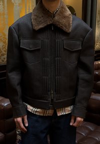 Veste gris foncé avec un col en fausse fourrure marron, des plis à l'avant et deux poches poitrine. Portée sur une chemise à carreaux et associée à un jean foncé.