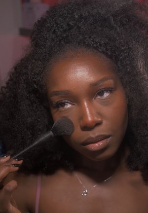 Joven con cabello rizado aplicándose maquillaje con una brocha en la mejilla, enfocada en su reflejo o espejo.